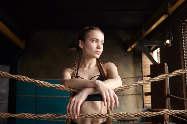 Attractive muscular young woman posing indoors in boxing ring, having rest after intensive workout