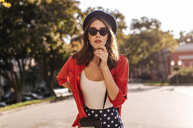 Attractive pale girl in French beret black sunglasses white top and shirt posing outdoors and touching face Warm and sunny weather