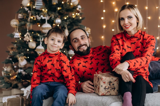 Attractive parents and their little son in red sweaters have fun opening presents before a Christmas