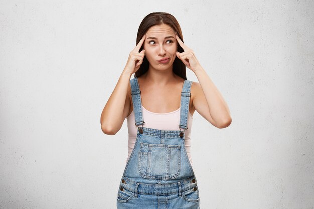 Attractive pensive woman dressed casually, keeping fingers on temples, curving lips and looking aside thinking over something important, trying to concentrate, isolated