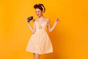 Free photo attractive pinup girl in dress holding camera. studio shot of smiling female photographer isolated on yellow space.