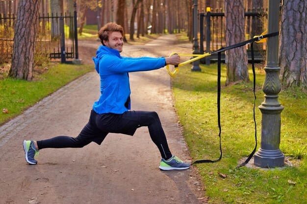 Attractive positive male in a blue raincoat exercising in a park with trx fitness strips.