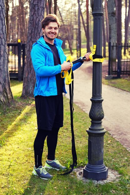 Attractive positive male in a blue raincoat exercising in a park with trx fitness strips.