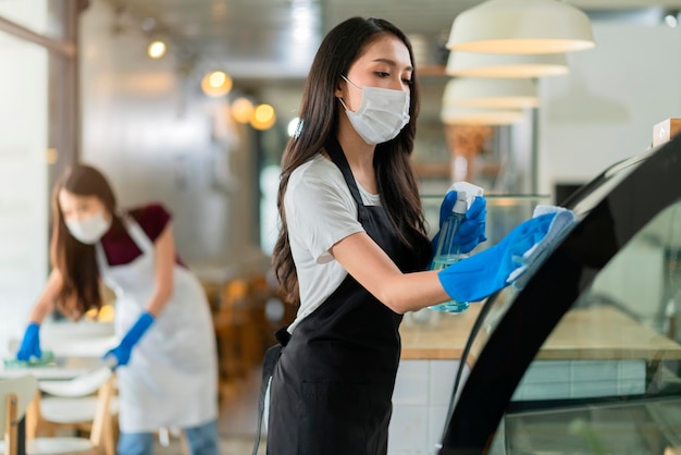 Free photo attractive positive young asian woman staff in uniform apron spraying detergent or alcohol on counter product cabinet wiping it with soft cloth when disinfecting cafe in ther morning safe and clean