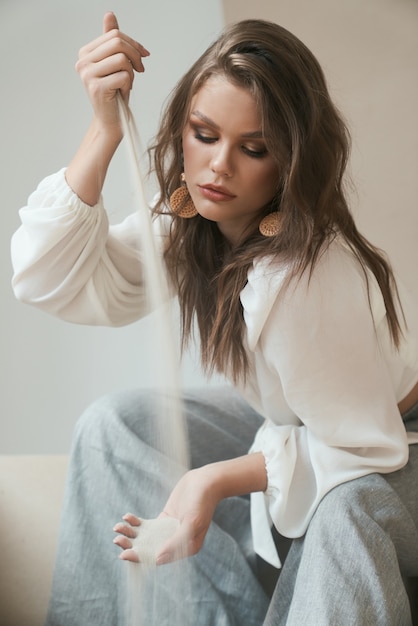 Attractive professional model with trendy make up and hairstyle looks very stylish in white blouse and grey trousers while pouring sea sand from one hand to another. Fashion looks. Modeling concept