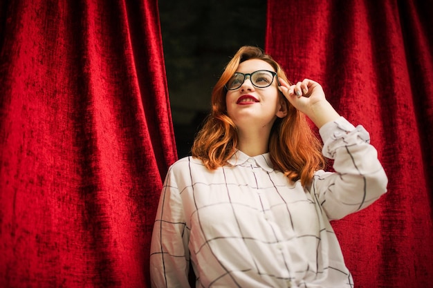 Attractive redhaired woman in eyeglasses wear on white blouse posing at arch of open red curtains