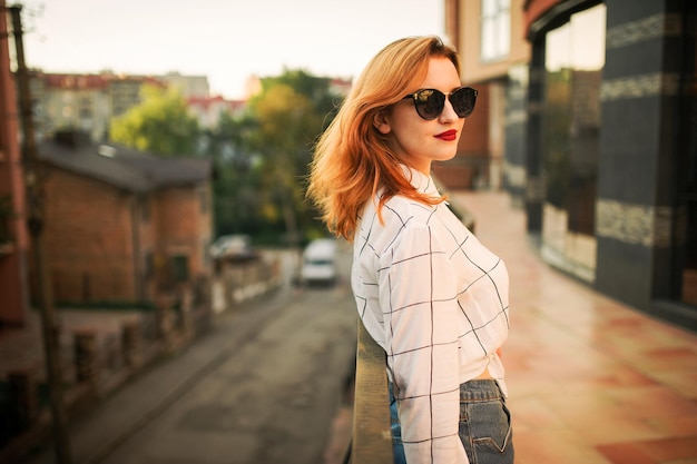 Attractive redhaired woman in sunglasses wear on white blouse posing at street against modern building