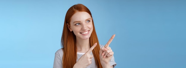 Free Photo attractive redhead female model pure clean skin ginger long hair pointing looking left intrigued smiling satisfied indicating interesting advertisement intrigued using new product blue background