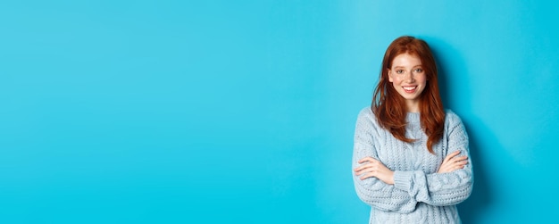 Free photo attractive redhead girl in sweater smiling and staring at camera standing confident against blue bac