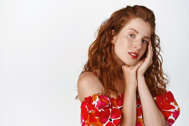 Attractive redhead woman touch her face looking coquettish at camera wearing floral dress standing over white background