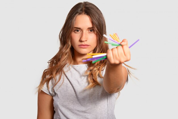 Attractive serious female holds crumples plastic straws in hand, demonstraes her strong feeling of living on clean planet