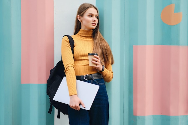 Attractive serious student girl with laptop and coffee to go looking away over colorful background outdoor