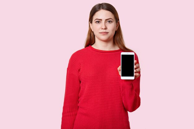 Attractive serious young European teenage girl with long hair, dressed in red loose jumper, holds modern cell phone with black empty