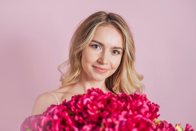 Free photo an attractive smiling blonde young woman with red flower bouquet against pink background