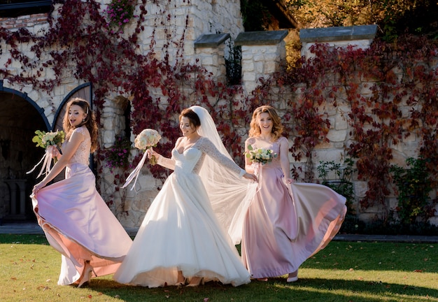 Free photo attractive smiling bride and bridesmaids are dancing and having fun in front of stone building covered with red ivy