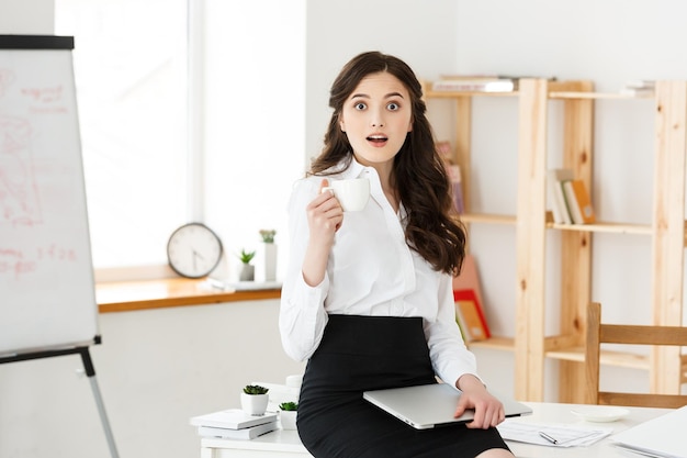 Attractive smiling business woman sitting at office desk holding a cup of coffee she is relaxing and