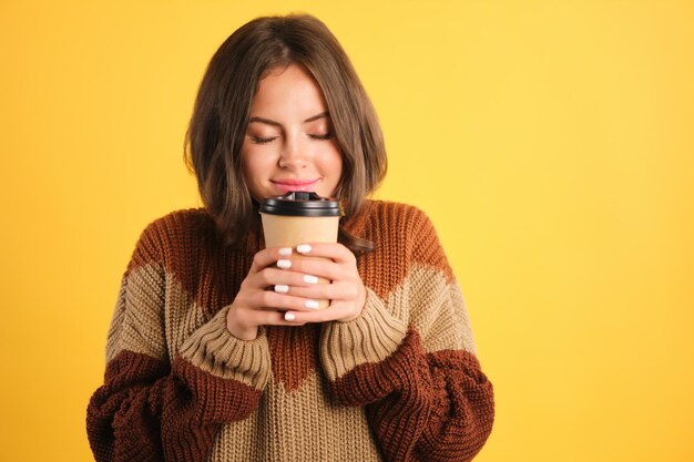 Attractive smiling girl in cozy sweater dreamily smelling coffee over yellow background