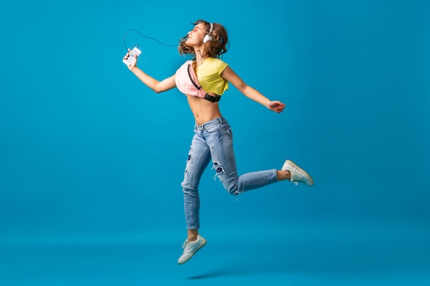 Free photo attractive smiling happy woman dancing listening to music in headphones dressed in hipster stylish outfit isolated on blue studio background, wearing colorful clothes