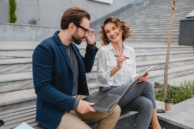 Free photo attractive smiling man and woman talking sitting on bench in urban city center, making notes