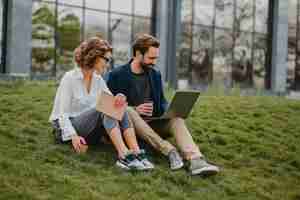 Free photo attractive smiling man and woman talking sitting on grass in urban park, making notes