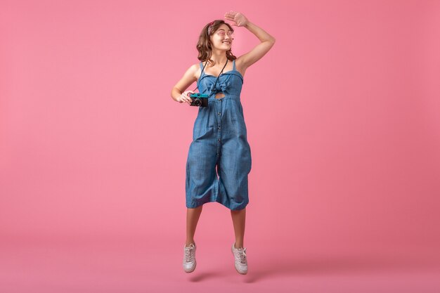 Attractive smiling woman jumping active taking photo on vintage camera wearing denim dress isolated on pink background