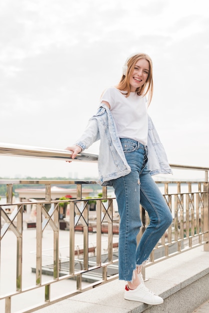 Free photo attractive smiling woman leaning on bridge railing and listening music