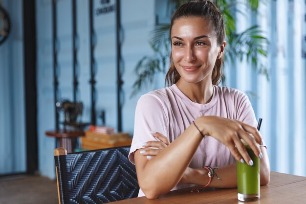 Attractive smiling woman on vacation, enjoying paradise resort, sits in a cafe using mobile phone and drinking healthy smoothie.