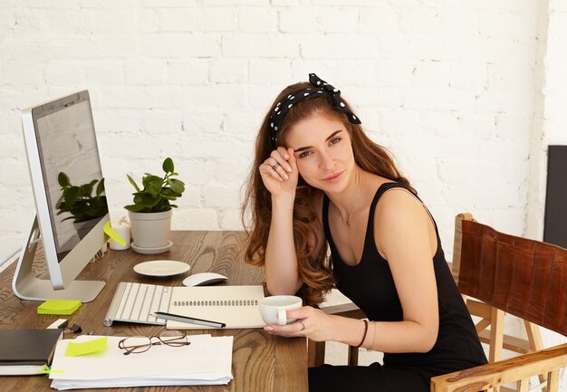 Attractive smiling young female freelancer enjoying cappuccino during coffee break while working remotely at her home office, using high-speed internet connection on copy space screen computer