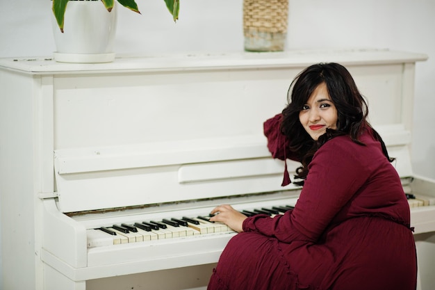 Free photo attractive south asian woman in deep red gown dress posed at room and play on piano