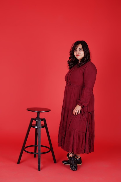 Free photo attractive south asian woman in deep red gown dress posed at studio on pink background sitting on chair