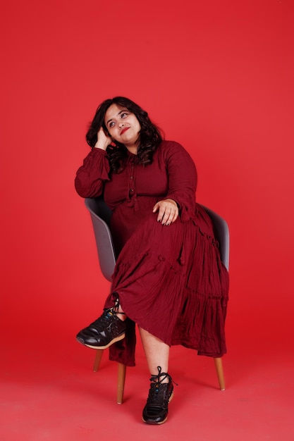 Attractive south asian woman in deep red gown dress posed at studio on pink background sitting on chair