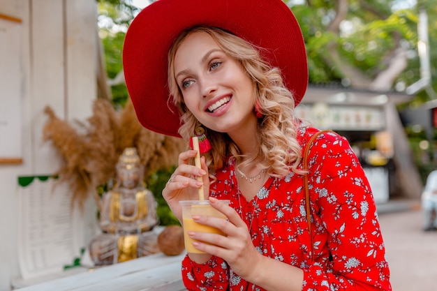 Attractive stylish blond smiling woman in straw red hat and blouse summer fashion outfit drinking natural fruit cocktail smoothie