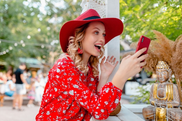 Attractive stylish blond smiling woman in straw red hat and blouse summer fashion outfit holding using smart phone  cafe