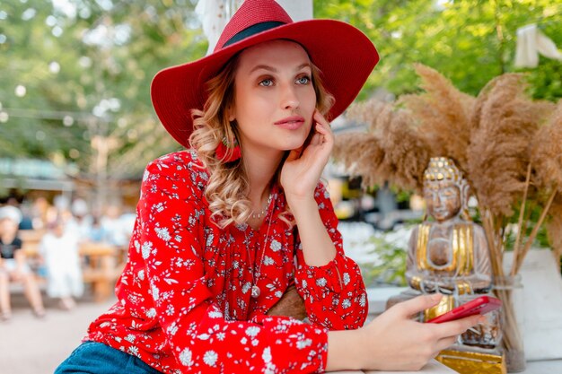 Attractive stylish blond smiling woman in straw red hat and blouse summer fashion outfit holding using smart phone  cafe