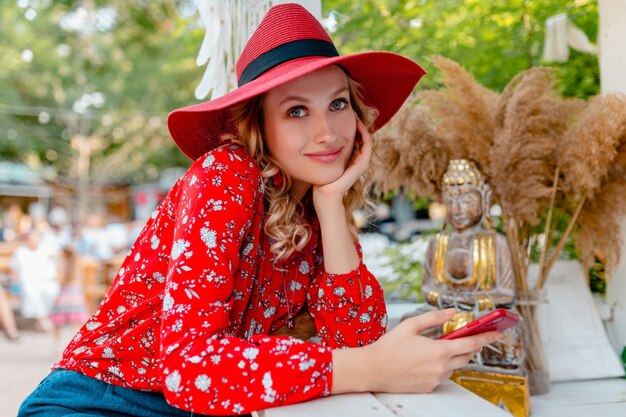 Attractive stylish blond smiling woman in straw red hat and blouse summer fashion outfit holding using smart phone  cafe