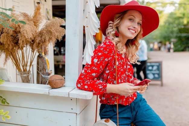 Free Photo attractive stylish blond smiling woman in straw red hat and blouse summer fashion outfit holding using smart phone  cafe