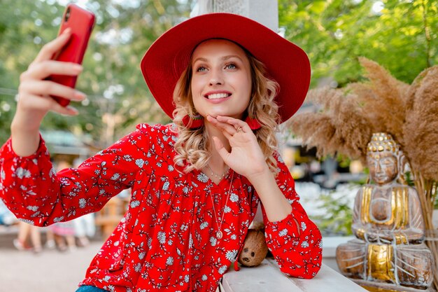 Attractive stylish blond smiling woman in straw red hat and blouse summer fashion outfit taking selfie photo on camera smart phone  cafe