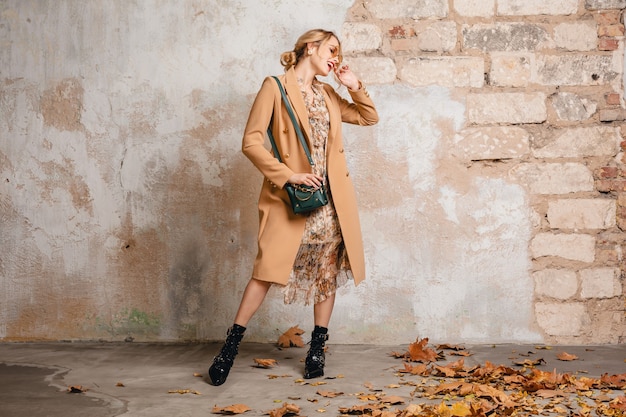 Free photo attractive stylish blonde woman in beige coat walking in street against vintage wall