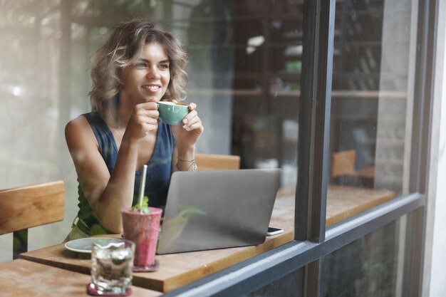 Attractive stylish female entrepreneur working outside office sit cafe look outside window passersby dreamy smiling holding cup coffee enjoy momentum working laptop prepare project