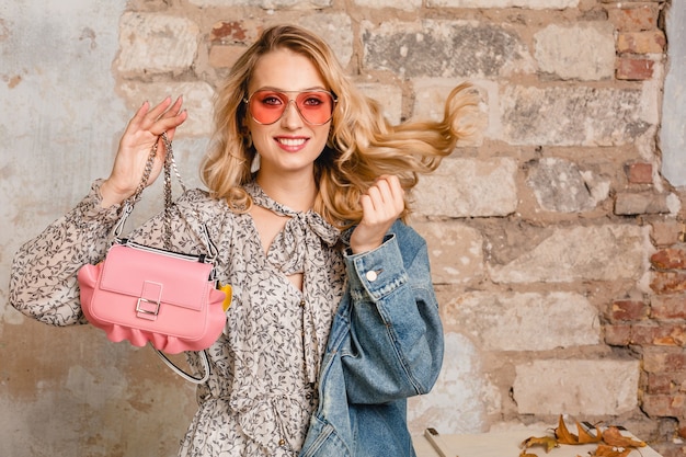 Attractive stylish smiling blonde woman in jeans jacket walking against wall in street