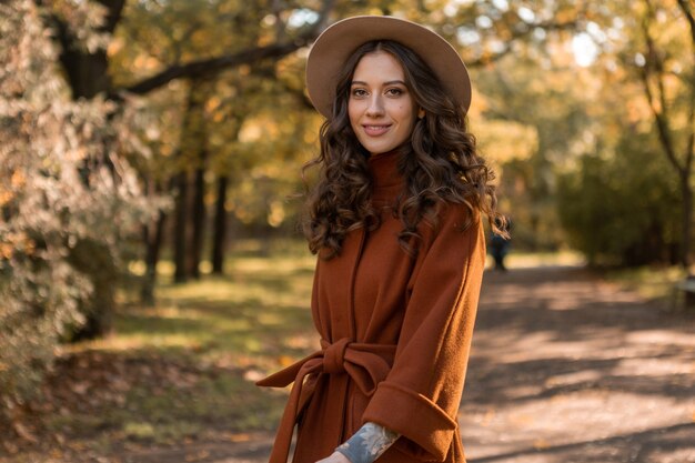 Attractive stylish smiling skinny woman with curly hair walking in park dressed in warm brown coat, autumn trendy fashion street style