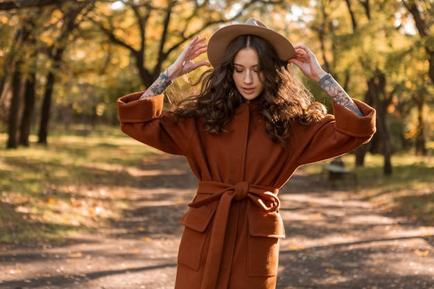 Free photo attractive stylish smiling skinny woman with curly hair walking in park dressed in warm brown coat, autumn trendy fashion street style