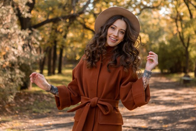 Attractive stylish smiling skinny woman with curly hair walking in park dressed in warm brown coat, autumn trendy fashion street style