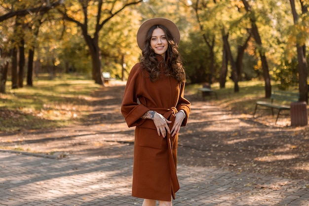 Attractive stylish smiling skinny woman with curly hair walking in park dressed in warm brown coat, autumn trendy fashion street style