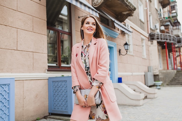 Attractive stylish smiling woman walking city street in pink coat, listening to music on earphones