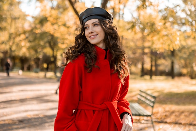 Attractive stylish smiling woman with curly hair walking in park dressed in warm red coat autumn trendy fashion, street style, wearing beret hat