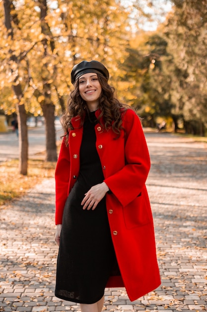 Attractive stylish smiling woman with curly hair walking in park dressed in warm red coat autumn trendy fashion, street style, wearing beret hat