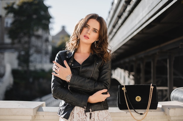 Attractive stylish woman posing in street in fashionable outfit, suede handbag, wearing black leather jacket and white lace dress, spring autumn style