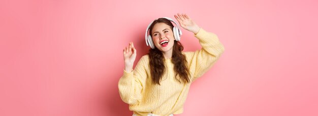 Free Photo attractive stylish woman singing favorite song listening to music in wireless headphones smiling and dancing standing over pink background