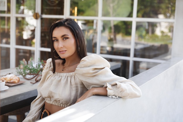 Free photo attractive stylish woman sit outdoor cafe table eating croissant and drinking coffee sexy female model look camera lean on fence as enjoying meal in coffeeshop restaurant diner